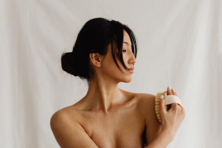 Una mujer con el pelo oscuro recogido en un moño bajo está de pie sobre un fondo blanco, sosteniendo un cepillo corporal en el hombro y mirando a un lado. Parece estar practicando el autocuidado o el cepillado en seco, tal vez relajándose después de leer información técnica.