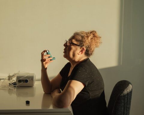 Una mujer mayor con gafas está sentada en una mesa, sosteniendo un inhalador cerca de la boca. Bañada por la cálida luz del sol, parece pensativa, mientras a su lado descansa un equipo médico y un pequeño tapón de botella, tal vez leyendo información tecnológica actualizada en su dispositivo.