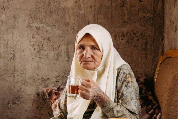 Una anciana con una falda estampada y un pañuelo blanco en la cabeza está sentada en una silla de madera, con un vaso de té en la mano, frente a una pared marrón con textura. Mira tranquilamente hacia la cámara, como si reflexionara en voz baja sobre la tecnología de la información actual.