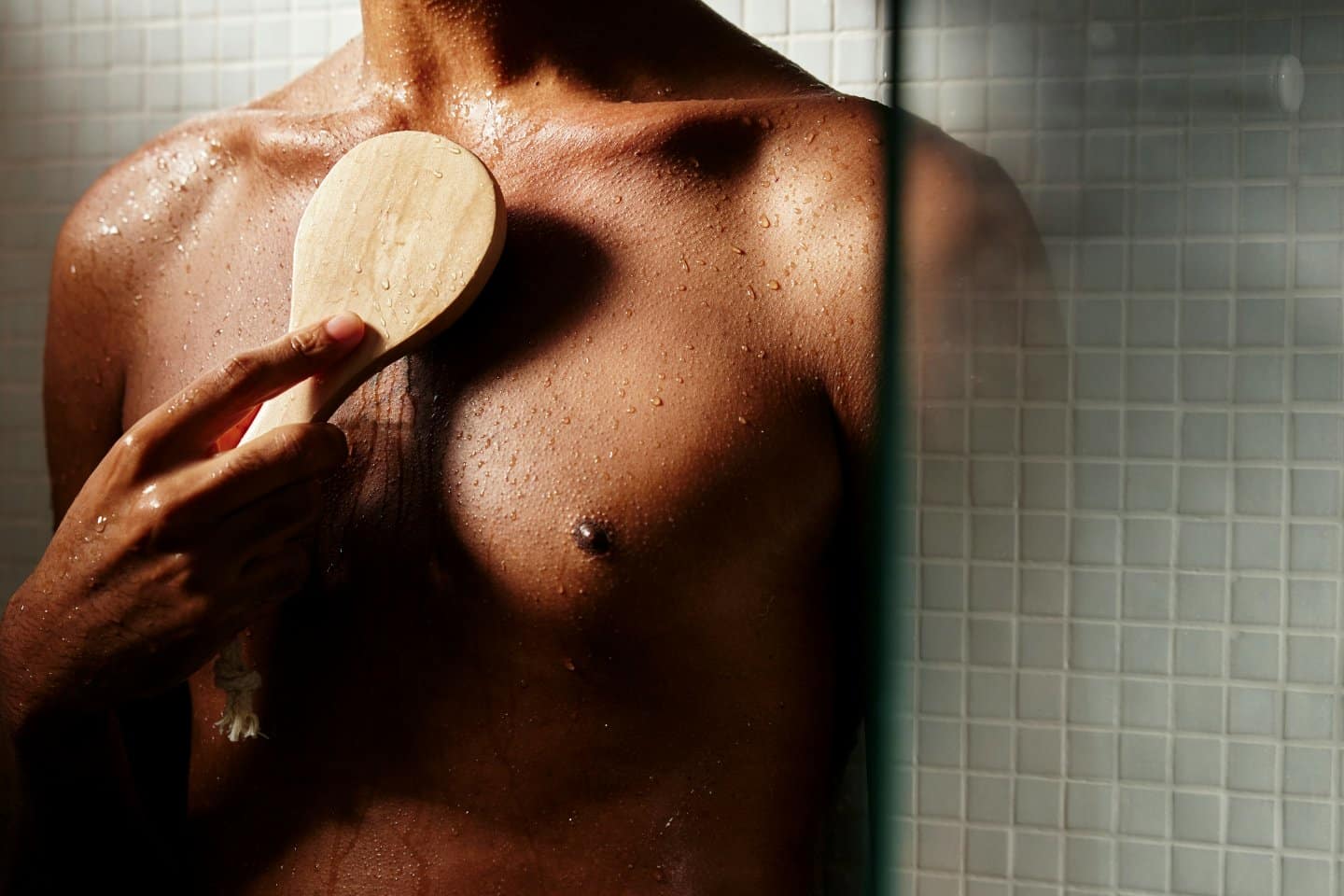 Una persona en la ducha utiliza un cepillo de madera para frotar la parte superior del pecho, con gotas de agua sobre la piel, una pared de azulejos en el fondo, tal vez tomando un descanso de ponerse al día en información tech durante su rutina.